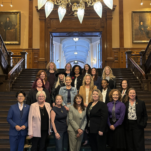 Group photo of approximately 18 people standing on the grand staircase inside Ontario's Legislative Building at Queen's Park in Toronto. The group is arranged in several rows beneath an ornate chandelier, with wood-paneled walls, historic portraits, and an arched corridor visible in the background.